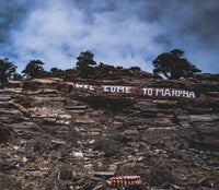 Large wooden sign with 'Welcome to Marpha' on a rocky cliff with trees and blue sky.