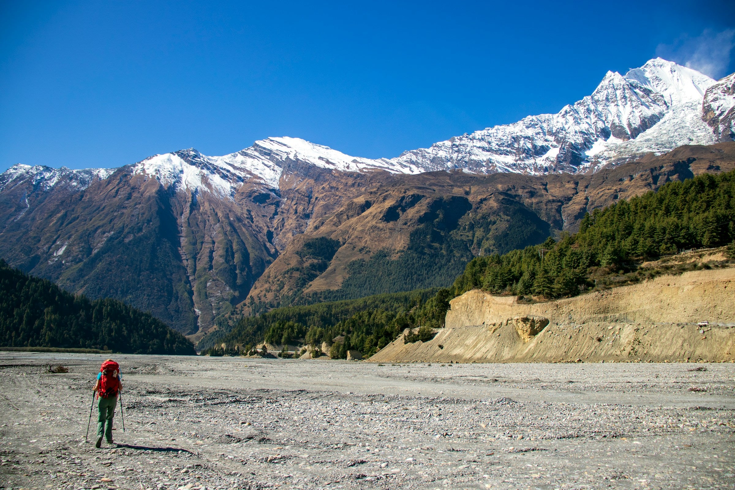Person with a red backpack walking on a gravel path with snow-capped mountains in the background