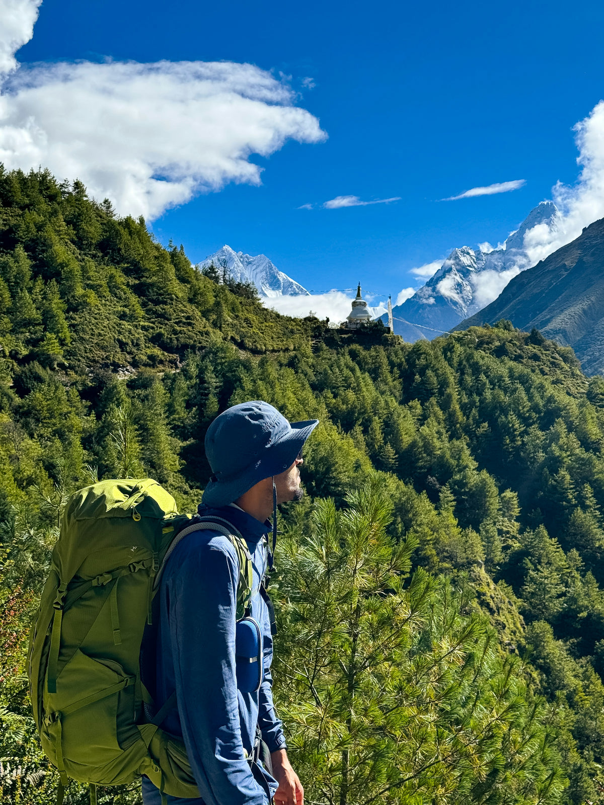 Person with a green backpack on a mountain trail, with mountains and clouds beyond