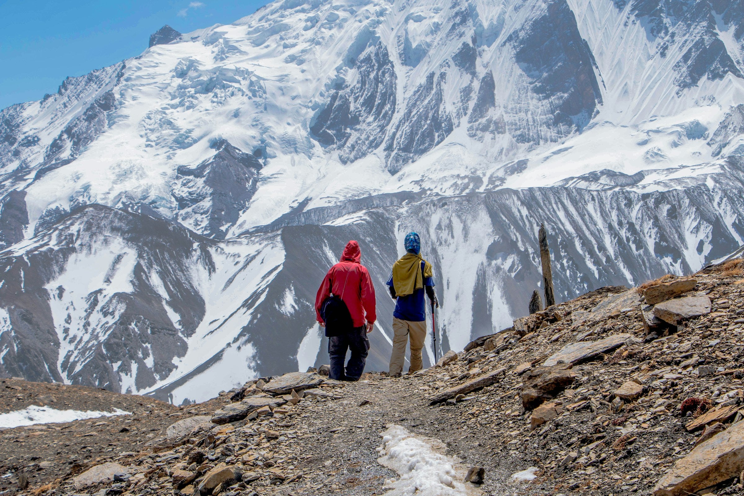 Two hikers on a mountain trail with snow-capped peaks in the background