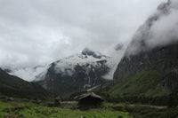 Mountain landscape with a small hut and snow-capped peaks under cloudy skies.