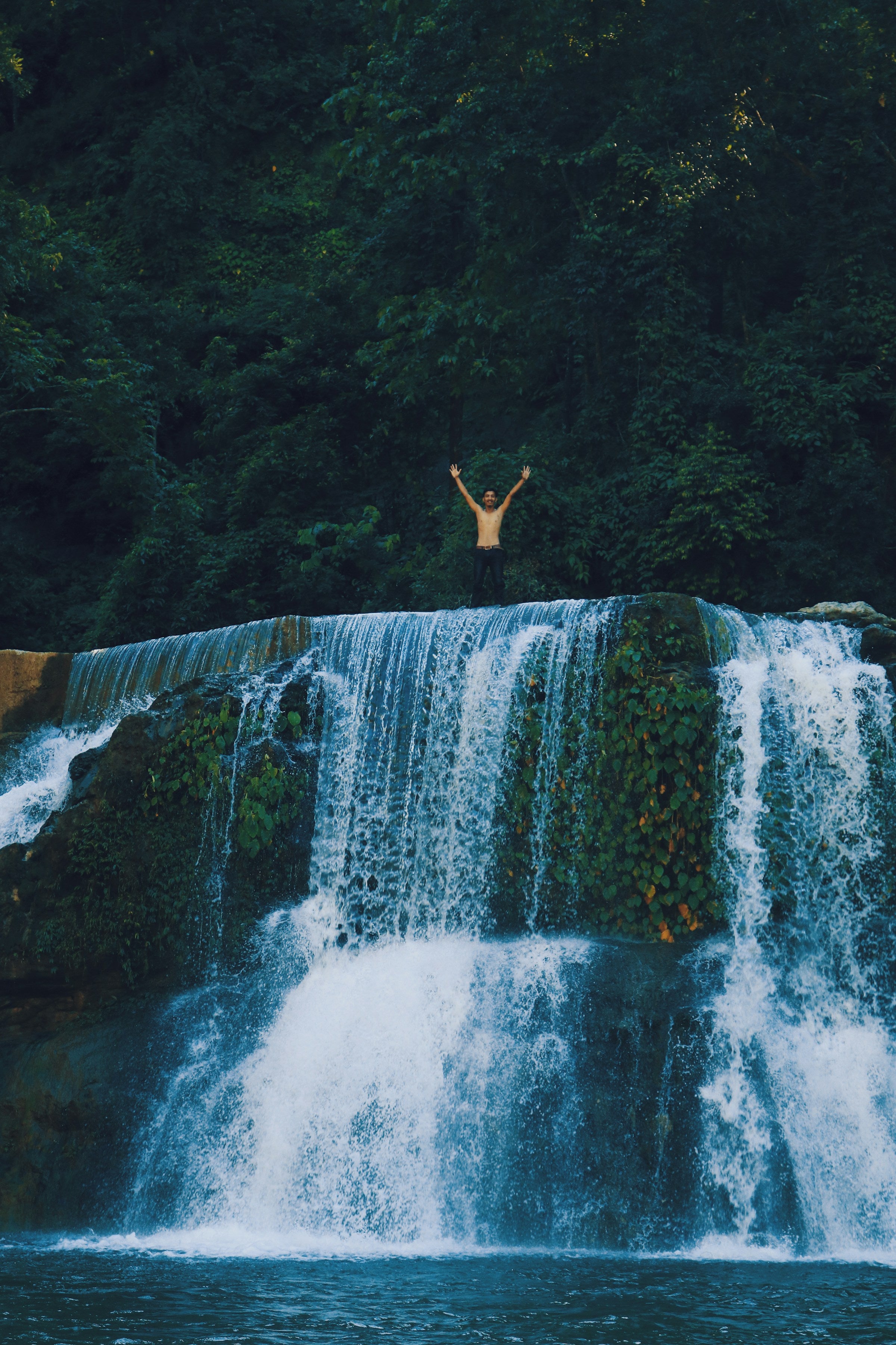 Person standing at the base of a waterfall with arms raised, surrounded by lush greenery.